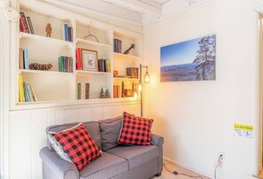 Una sala de estar acogedora con un sofá gris, cojines de cuadros rojos y negros, y una estantería llena de libros y objetos decorativos. En la pared, hay un cuadro enmarcado de un árbol, y se observa una señal de precaución.