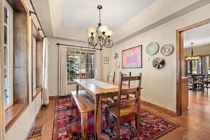 Un coin repas avec une table en bois, des bancs et un lustre. Des assiettes décoratives ornent le mur et un tapis à motifs recouvre le sol.