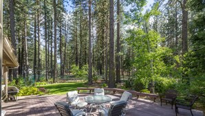 A wooden deck with a glass-top table and chairs, surrounded by tall trees.