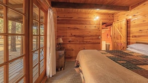 A wooden cabin bedroom with a bed, a nightstand, a lamp, and a window with curtains. - Forest Cabin 4 Cowboys Dream (Payson)