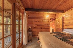Un dormitorio de cabaña de madera con una cama, una mesita de noche, una lámpara y una ventana con cortinas.