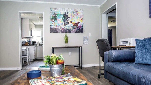 A living room with a dark blue sofa, a coffee table with a Star Wars magazine, a colorful abstract painting, and a kitchen visible through an open doorway. - "Rebel Base" in the  Heart of Fayetteville (Fayetteville)