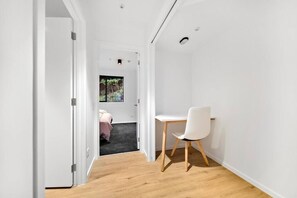 A narrow hallway with a wooden floor leading to a bedroom with a bed and a window. A white desk and chair are positioned in the hallway. - Stylish Cosy 1 Bedroom Apartment, minutes walk to town (Queenstown)