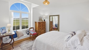 A bedroom with a large bed, a dresser, a chair, a desk with a lamp, a mirror, and a large window with a view of the outdoors. - Spectacular beachfront with epic views, beautiful lit-up deck for sunset dining! (Lubec)