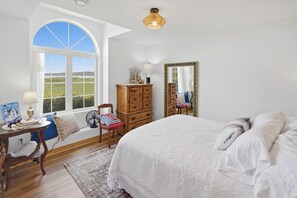 A bedroom with a large bed, a dresser, a chair, a desk with a lamp, a mirror, and a large window with a view of the outdoors.