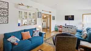 A living room with a blue sofa, a wooden coffee table, a television, and a kitchen area in the background. - Spectacular beachfront with epic views, beautiful lit-up deck for sunset dining! (Lubec)