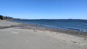 Una playa con cielo despejado, agua tranquila y una costa lejana con árboles y edificios.
