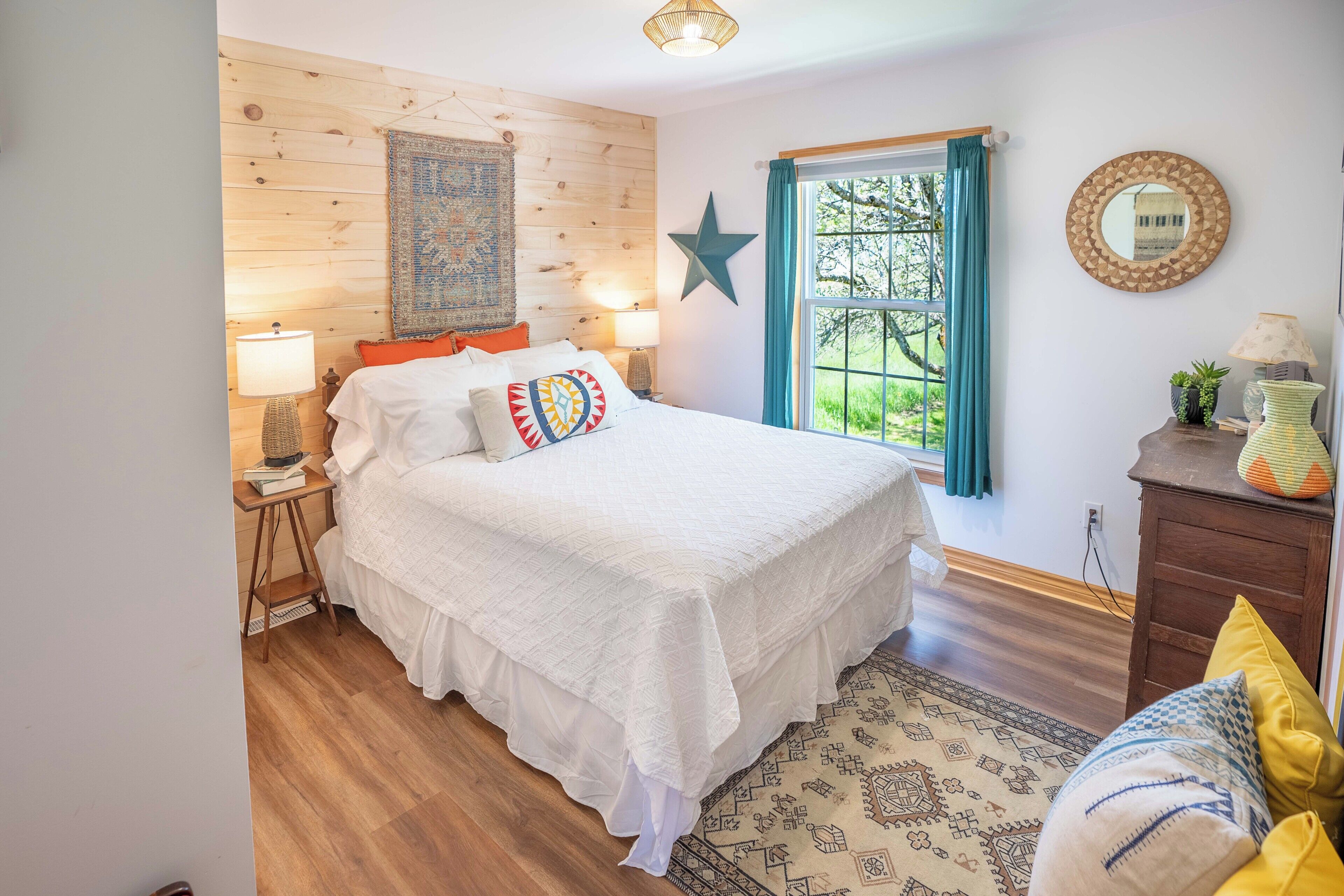 A bedroom with a wooden headboard, a star decoration, a window with curtains, a bedside table with a lamp, and a patterned rug.