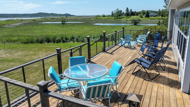 A deck with outdoor furniture, a table, and chairs, overlooking a natural landscape with water bodies and greenery. - Spectacular beachfront with epic views, beautiful lit-up deck for sunset dining! (Lubec)