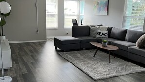 Ein modernes Wohnzimmer mit einem grauen Ecksofa, einem kleinen Couchtisch und einem grauen Shaggy-Teppich. An der Wand befindet sich eine weiße Klimaanlage und eine Stehlampe.