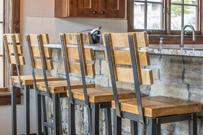 A kitchen with four wooden chairs with metal frames, a stone wall, and wooden cabinets above a sink.