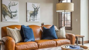 Un divano in pelle marrone con cuscini blu e a righe, un tavolino da caffè in legno con una rivista, e due quadri raffiguranti cervi appesi al muro.