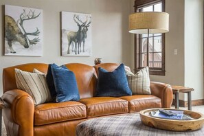 Ein braunes Ledersofa mit blau gestreiften Kissen, ein Couchtisch aus Holz mit einer Zeitschrift und zwei Hirschbilder an der Wand.
