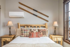 A bedroom with a wooden headboard, multiple patterned pillows, bedside lamps, and an air conditioning unit on the wall.