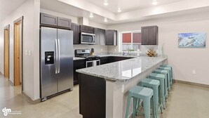 A modern kitchen with a stainless steel refrigerator, a microwave, and dark cabinets. There is a large island with a granite countertop and bar stools. - Upper Mesa Villa #9, Glacier Suites - Upper Mesa Villa #11 (Palmer)