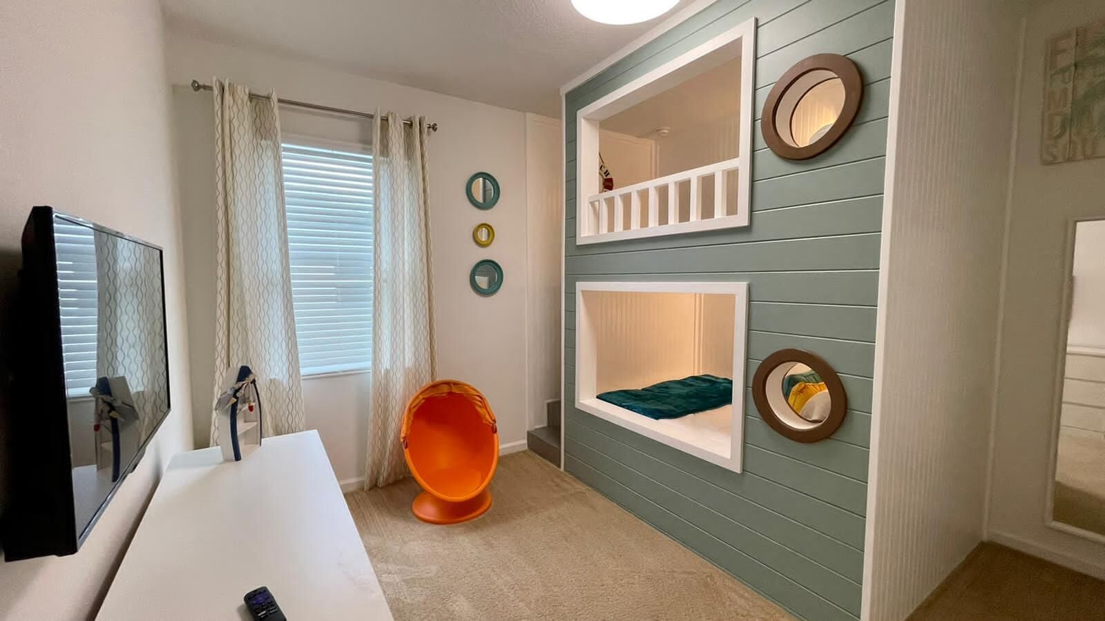 Une pièce avec un bureau blanc, une chaise, un lit recouvert d’une couverture verte et une fenêtre avec des rideaux.