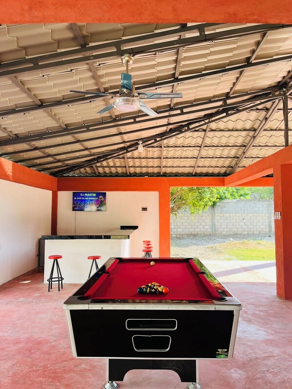A pool table in a covered area with a ceiling fan, a bar area, and a view of the outdoors.