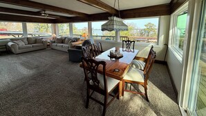 Amplia sala de estar con una mesa de comedor de madera, múltiples sillas y un conjunto de sofás. Amplias ventanas ofrecen vistas al exterior.