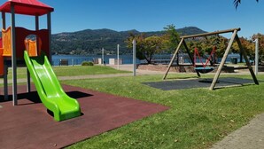 Un terrain de jeux avec un toboggan vert et des balançoires, le tout situé dans un cadre pittoresque, avec des collines en arrière-plan et un plan d'eau.