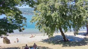 Une plage où des gens se prélassent au soleil et se reposent à l’ombre des arbres, avec une vue sur un lac paisible et des collines en arrière-plan.