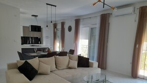 Un salon moderne avec un canapé d’angle beige, une table basse en verre et un coin repas avec une cuisine en arrière-plan.