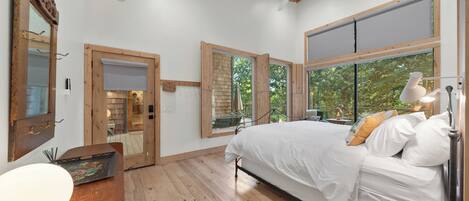 Quarto com cama, piso de madeira, ventilador de teto, janelas amplas e vista para o verde lá fora.