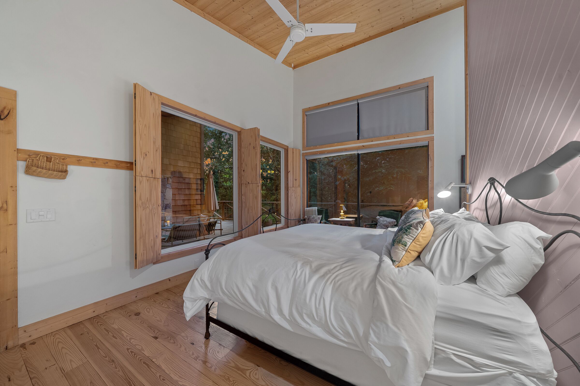 Una camera da letto con un letto, pavimento in legno, ventilatore a soffitto, ampie finestre e vista sull'esterno.