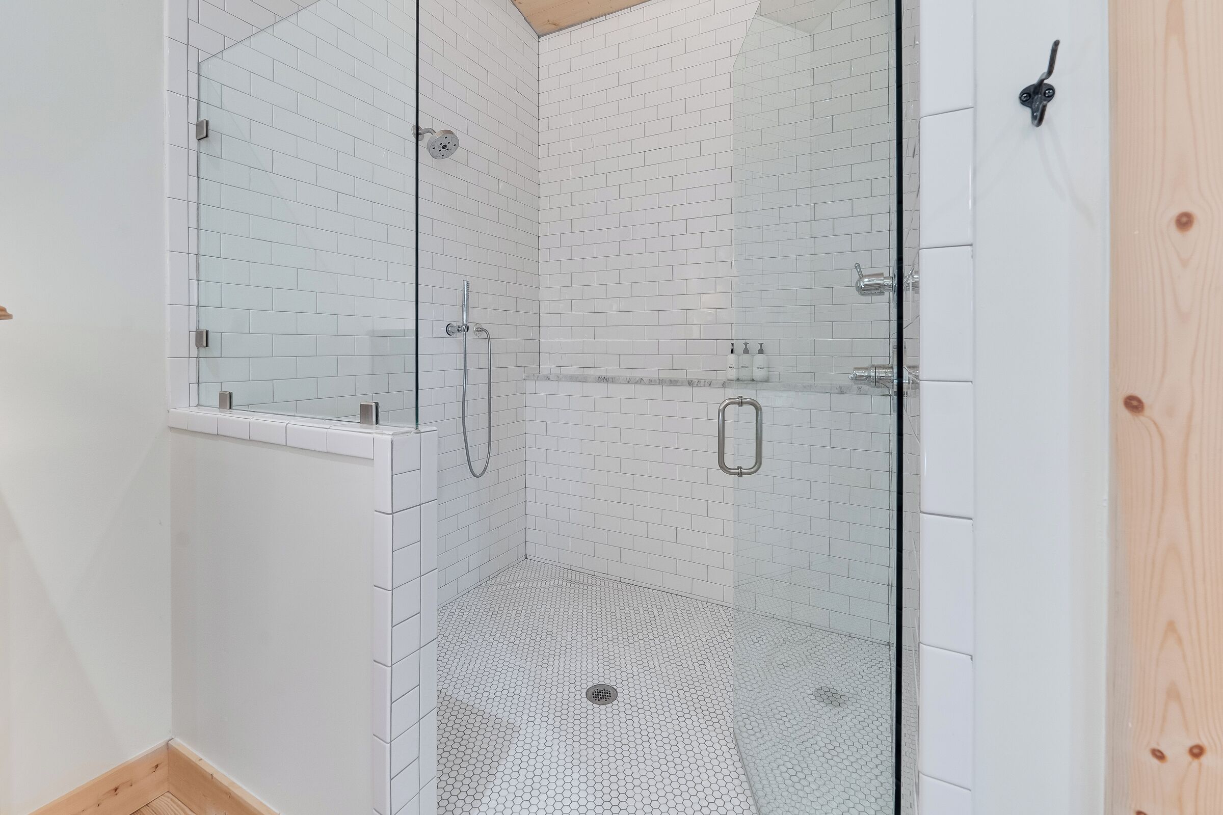 A glass-enclosed shower with white tiled walls and floor, a wooden door, and a metal showerhead.