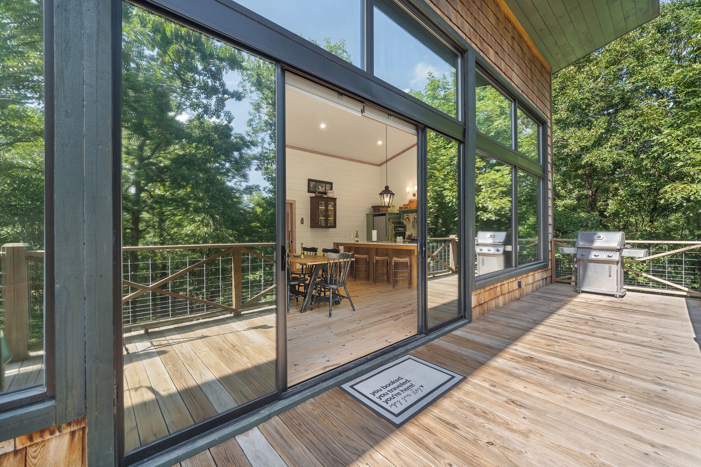 Une terrasse en bois avec une porte vitrée donnant sur un espace intérieur comprenant un coin repas, une cuisine et un barbecue.