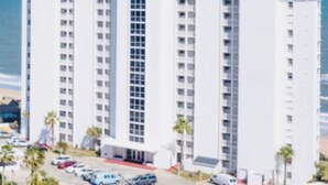 Un edificio de gran altura con varios pisos, un estacionamiento con autos y palmeras en primer plano.