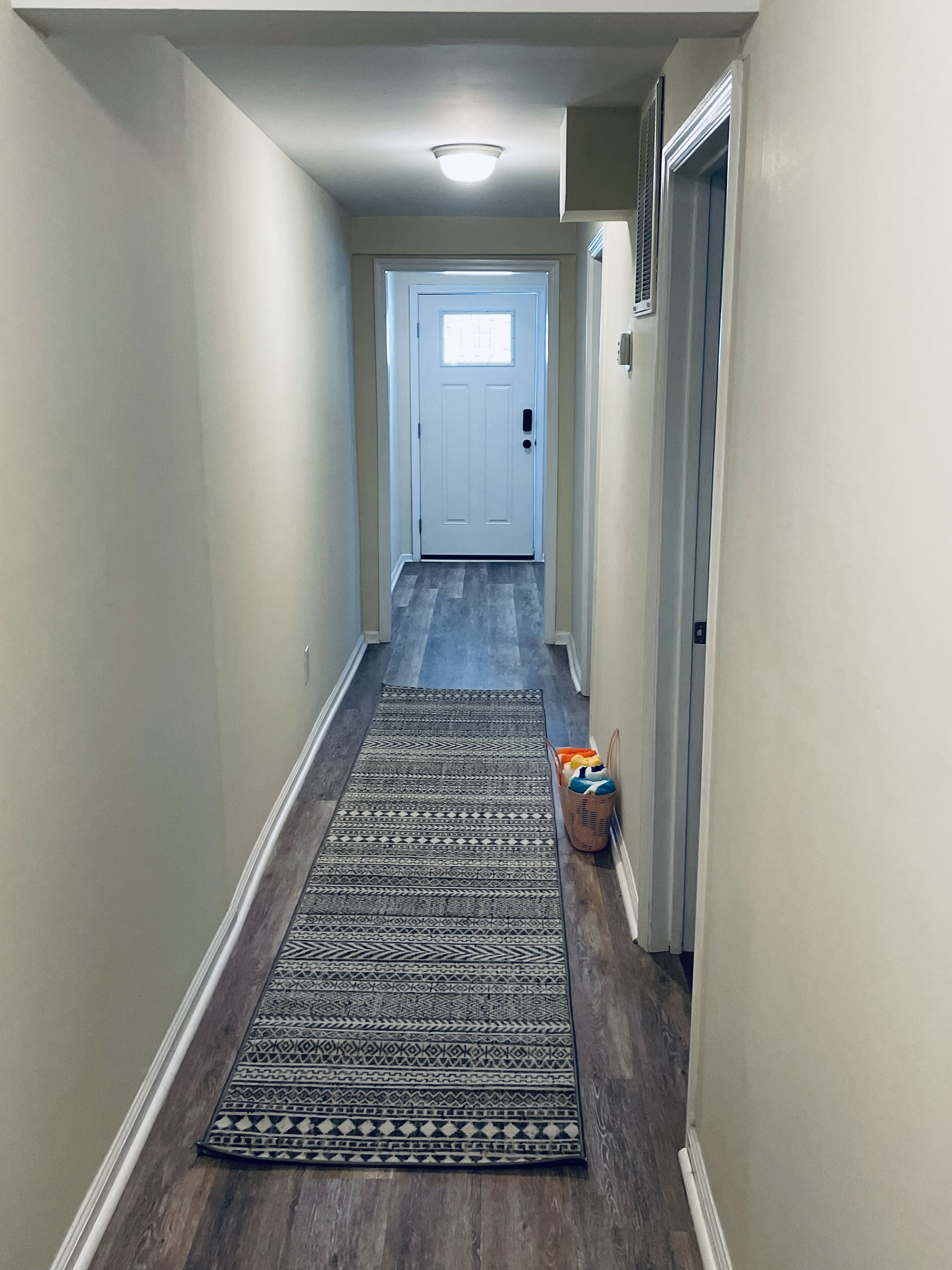 A narrow hallway with a patterned rug, a door at the end, and a basket with items on the floor.