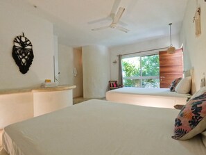 A bedroom with a bed, a bedside table, a window with a view of greenery, a ceiling fan, and a wall-mounted decorative item. - Rustik-inn (Playa del Carmen)