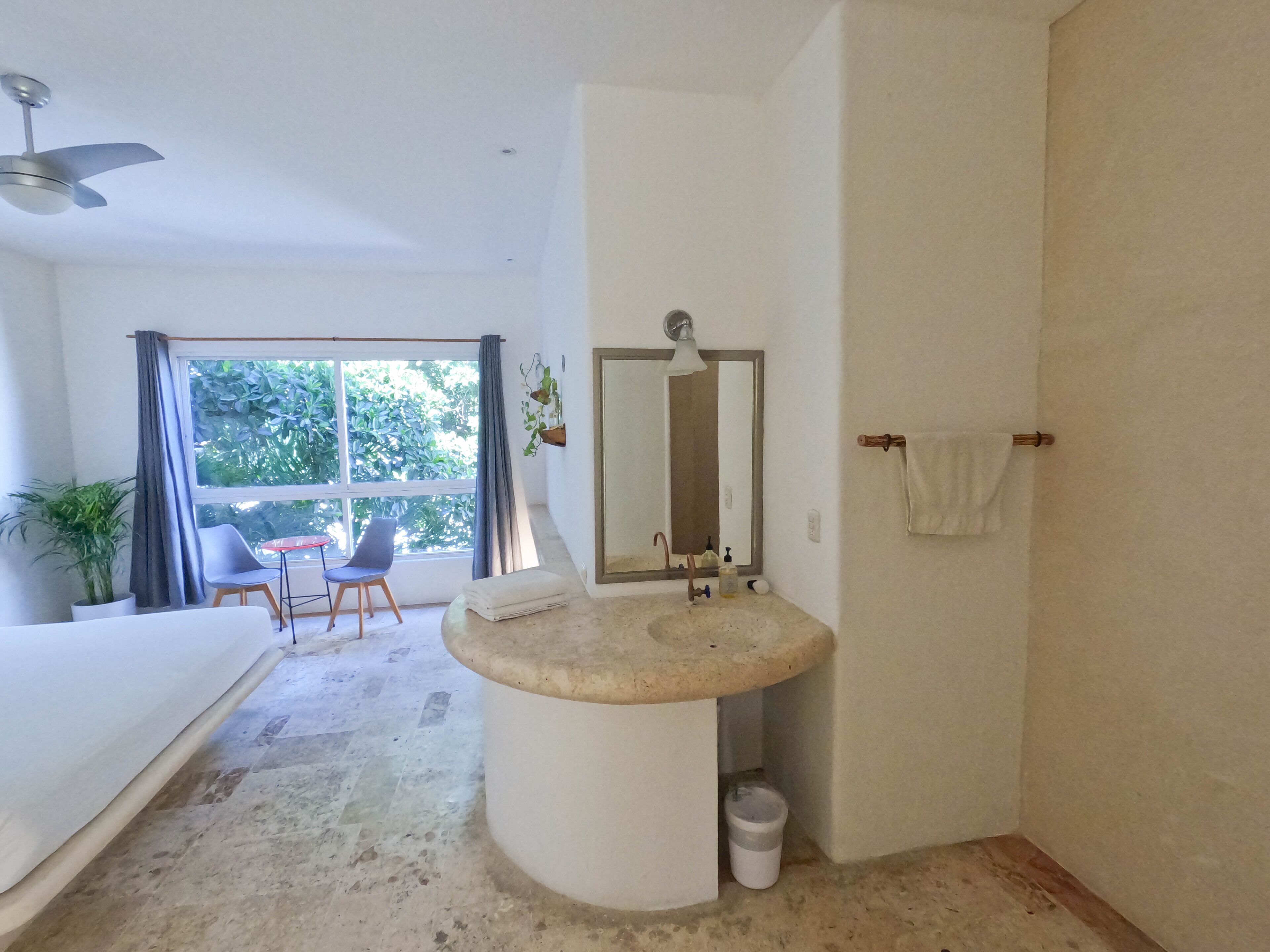 A bathroom with a round sink, a mirror, and a towel rack. There is a window with curtains and a plant outside.
