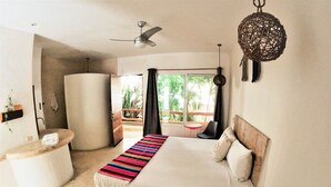 A bedroom with a bed, a bedside table, a chair, a ceiling fan, and a window with a view of greenery. - Rustik-inn (Playa del Carmen)