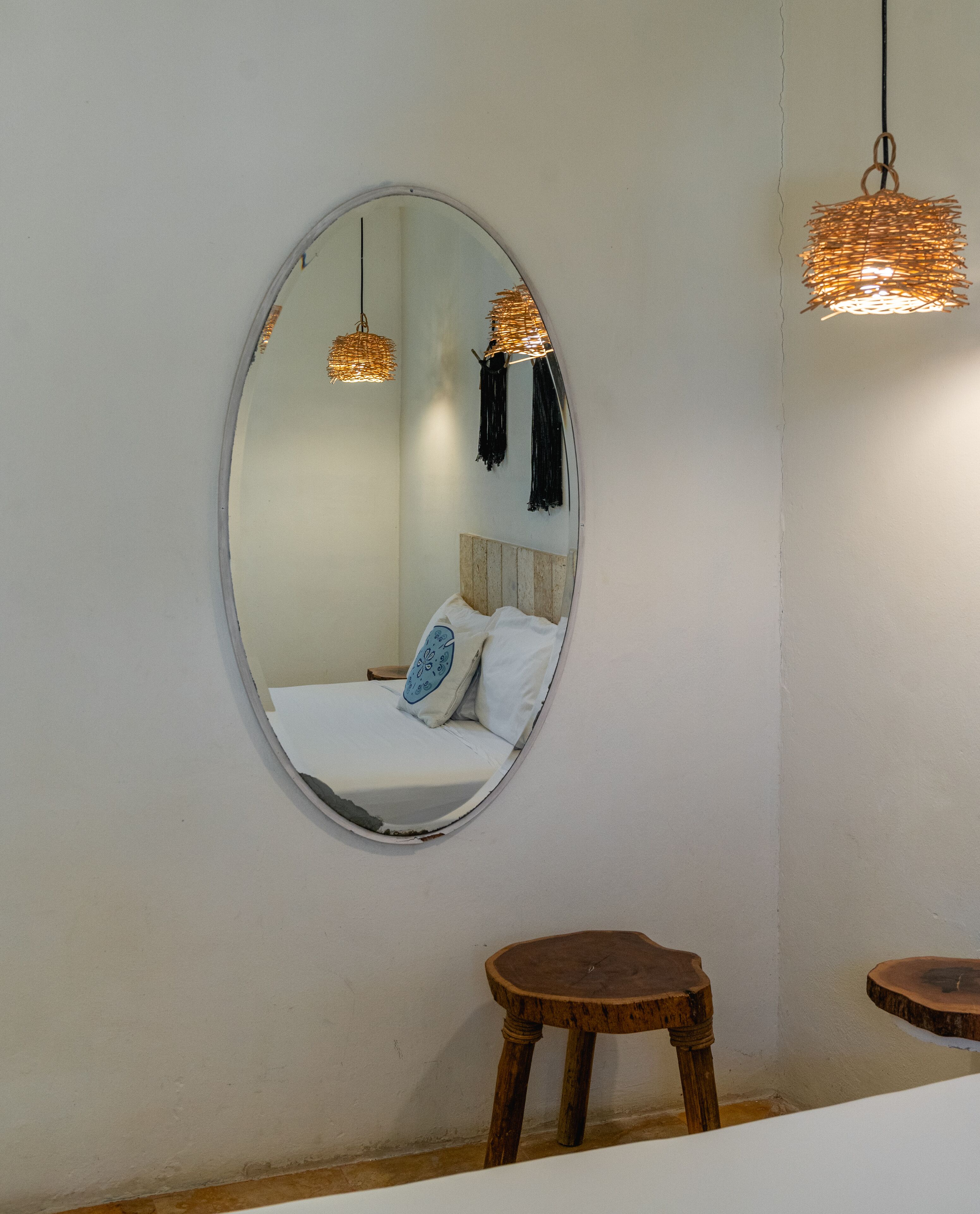 A room with a circular mirror on the wall, a bed with white bedding and patterned pillows, two wooden stools, and two hanging light fixtures.