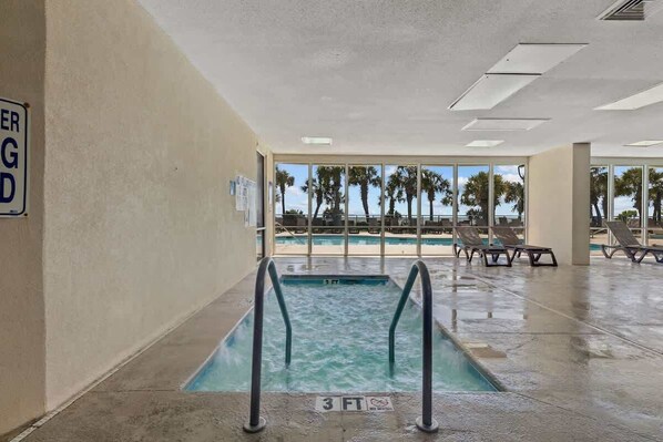 An indoor swimming pool with a 3 FT depth sign, lounge chairs, and a view of palm trees through large windows. - The Renewed Sunrise Beach Suite (Myrtle Beach)