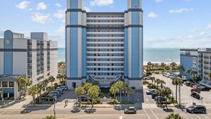 Un hotel con entrada principal, varios pisos y un estacionamiento con coches y palmeras.