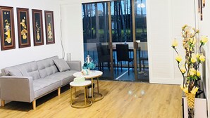 A living room with a grey sofa, a small round table with a teapot, and a vase with yellow flowers. There are four framed artworks on the wall, a ceiling fan, and a sliding glass door leading outside. - Hilo SunShine Rental.2bds1ba,common swimming pool. (Hilo)