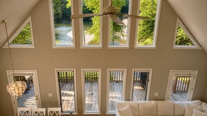 A spacious loft with a high ceiling, multiple windows offering views of a wooded area, a ceiling fan, and a cozy seating area. - Updated Lakefront Retreat, Fiber WiFi, Fire Pit, 15 Min to VIR (Leasburg)