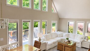 A spacious living area with a high ceiling, large windows, a white sectional sofa, a dining table, and a coffee table. - Updated Lakefront Retreat, Fiber WiFi, Fire Pit, 15 Min to VIR (Leasburg)