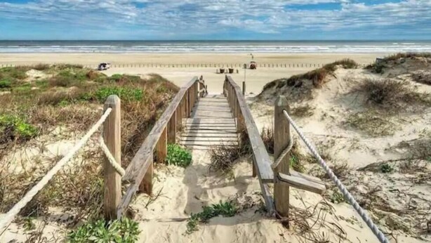 En träpromenad som leder till en strand med en sandstig och gräsklädda sanddyner.