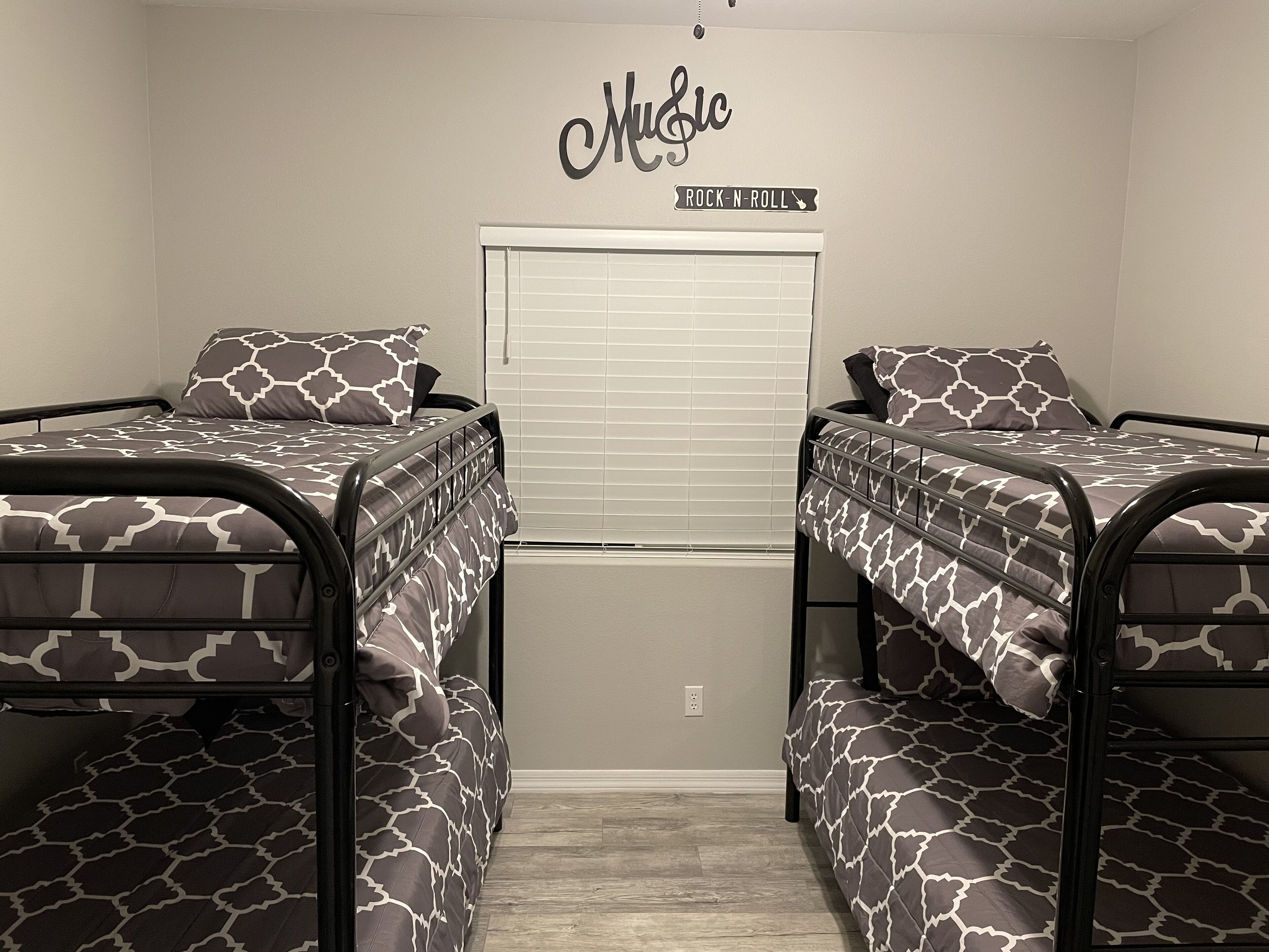 A room with two bunk beds, a window with blinds, a wall sign reading "Music", and a power outlet on the wall.