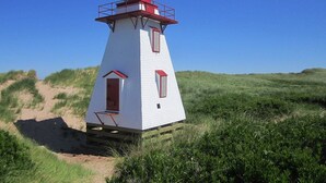 A white lighthouse with a red top, situated on a grassy dune with a clear blue sky in the background. - The Glowbug Cottage (Morell)