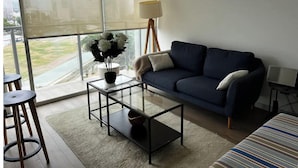 Ein modernes Wohnzimmer mit einem dunkelblauen Sofa, einem Glastisch und Blick auf eine Stadtlandschaft durch große Fenster.