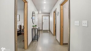A hallway with a bed, bedside table, and a mirror on the left. There is a door at the end of the hallway. - Upper Mesa Villa #11, Glacier Suites - Upper Mesa Villa #4 (Matanuska-Susitna)