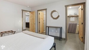 A bedroom with a bed, a bedside table, a wardrobe, and a mirror. There is a door leading to another room and a wall-mounted wreath. - Upper Mesa Villa #11, Glacier Suites - Upper Mesa Villa #4 (Matanuska-Susitna)