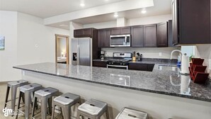 A modern kitchen with dark cabinets, stainless steel appliances, a granite countertop, and bar stools. - Upper Mesa Villa #11, Glacier Suites - Upper Mesa Villa #4 (Matanuska-Susitna)