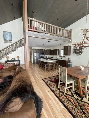 Un salón amplio con techo alto, una escalera de madera, un comedor con mesa y sillas de madera, y una cocina al fondo.