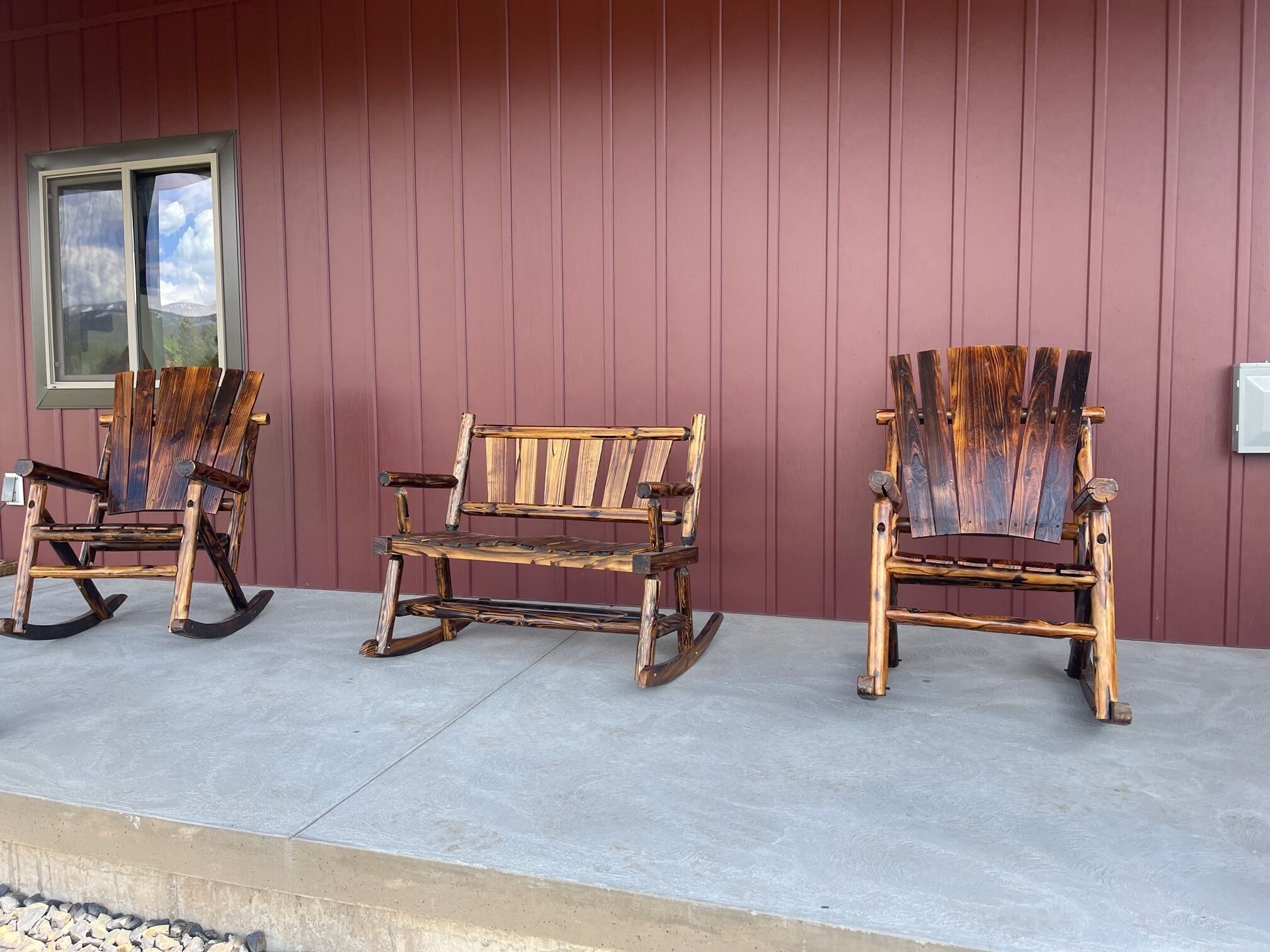 Three wooden rocking chairs and a bench on a concrete porch with a red wall and a window.