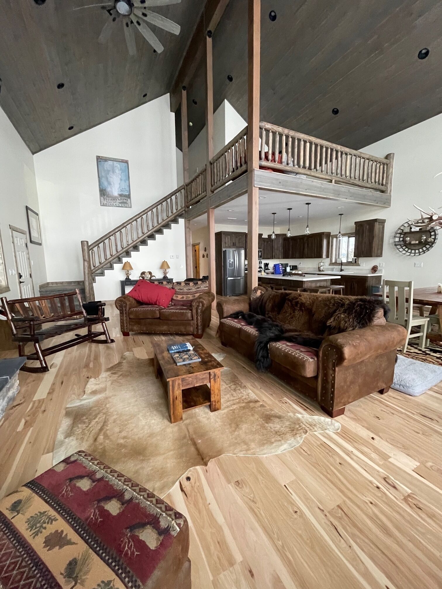 A spacious living area with a high ceiling, wooden flooring, a staircase to an upper level, and a kitchen area visible in the background.